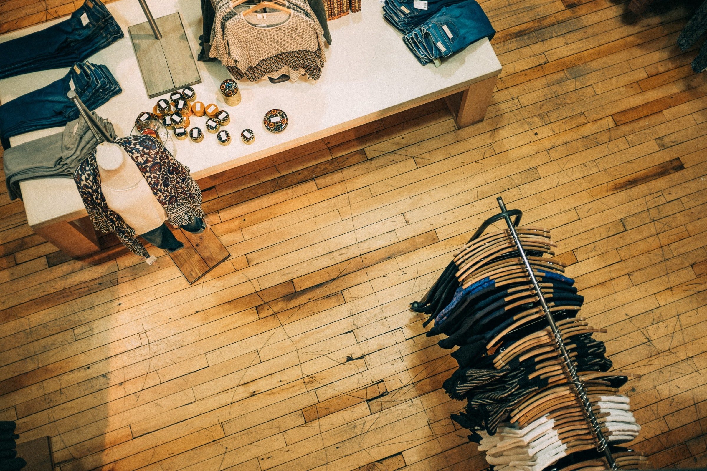 An overhead shot of a retail store interior showing light-colored hardwood floors. In the upper left, a white display table holds folded denim and patterned sweaters; in the lower right, a circular metal rack is filled with various garments on wooden hangers.